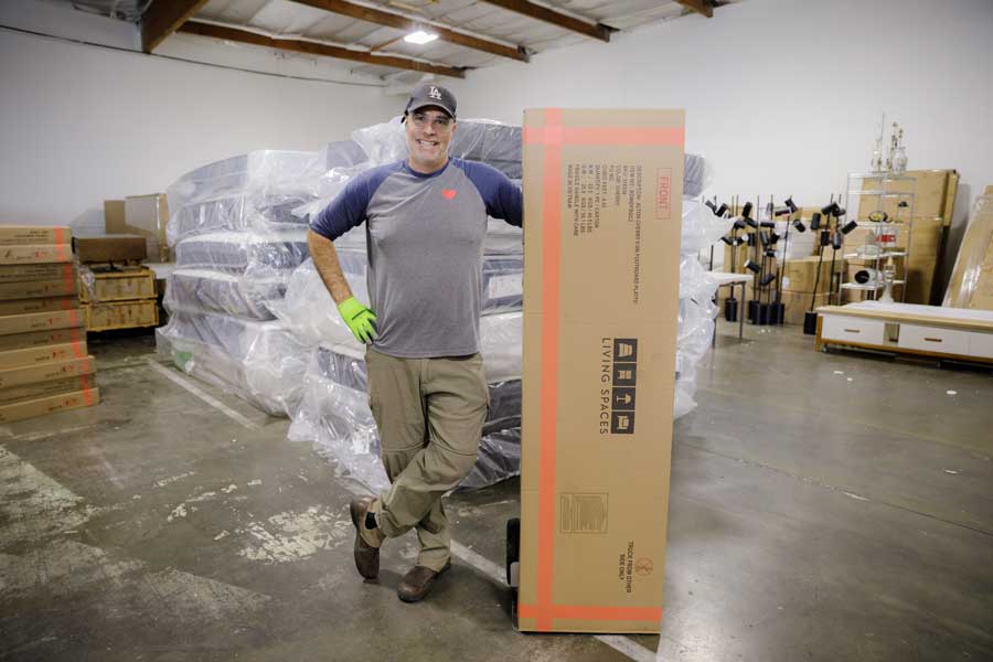 Alistair Campbell stands next a tall cardboard box while inside of a warehouse full of furniture.